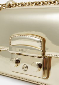 Kultainen metallinen käsilaukku, jossa on sileä pinta, ketjuhihna ja suorakaiteen muotoinen läppä, jossa on kaiverrettu "FURLA"-teksti sekä pyöreitä metallikoristeita.