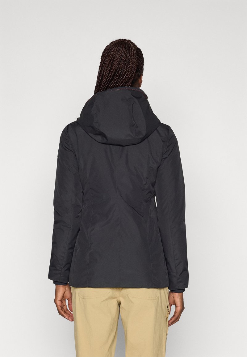 Frau mit geflochtenem Haar, die eine schwarze Kapuzenjacke und beigen Hosen trägt, von hinten vor einem schlichten hellen Hintergrund gezeigt.