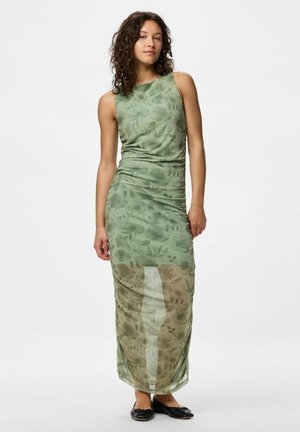 Femme portant une robe maxi fleurie verte sans manches, semi-transparente, avec des ballerines noires, debout devant un fond blanc uni.