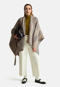 Károvaný pončo ve béžové barvě s modrými akcenty, kombinované se zeleným rolákem, bílými kalhotami širokého střihu a černou kabelkou. Má velký, pohodlný střih.