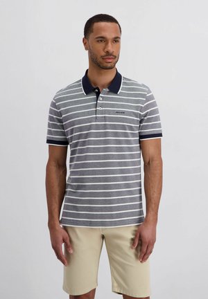 Mann trägt ein graues und weiß gestreiftes Poloshirt mit marineblauem Kragen und Bündchen sowie beigen Shorts, steht vor einem schlichten Hintergrund.