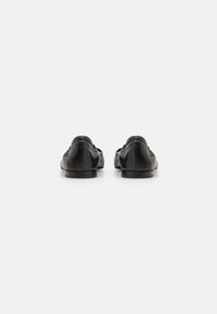 Zapatos negros de cuero tipo slip-on con una textura suave, puntera redondeada y tacón bajo, que muestran detalles de costura en la parte superior y un diseño minimalista.