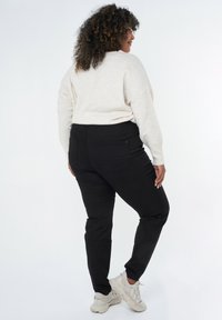 Pantalon noir extensible avec une texture lisse et un design ajusté, associé à un pull crème clair et des baskets beiges épaisses.