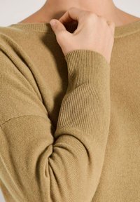 Eine Person passt den Halsausschnitt eines beigen, gerippten Pullovers an, der die detaillierte Stricktextur und die langen Ärmel zeigt.
