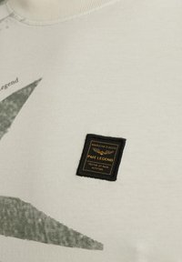 Sweat-shirt beige avec un design graphique vert foncé, col rond, et un patch carré noir avec un texte doré et un logo d'aigle.