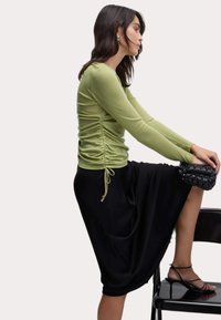 Top a maniche lunghe, di un verde chiaro, con arricciature laterali, abbinato a una gonna nera ampia. La modella è seduta su una sedia, mostrando sandali neri con cinturini.
