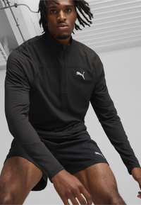 Schwarzes langärmliges Sportoberteil mit einem Reißverschlusskragen, ausgestattet mit einem weißen Puma-Logo auf der Brust, kombiniert mit schwarzen Shorts. Glatte Textur.