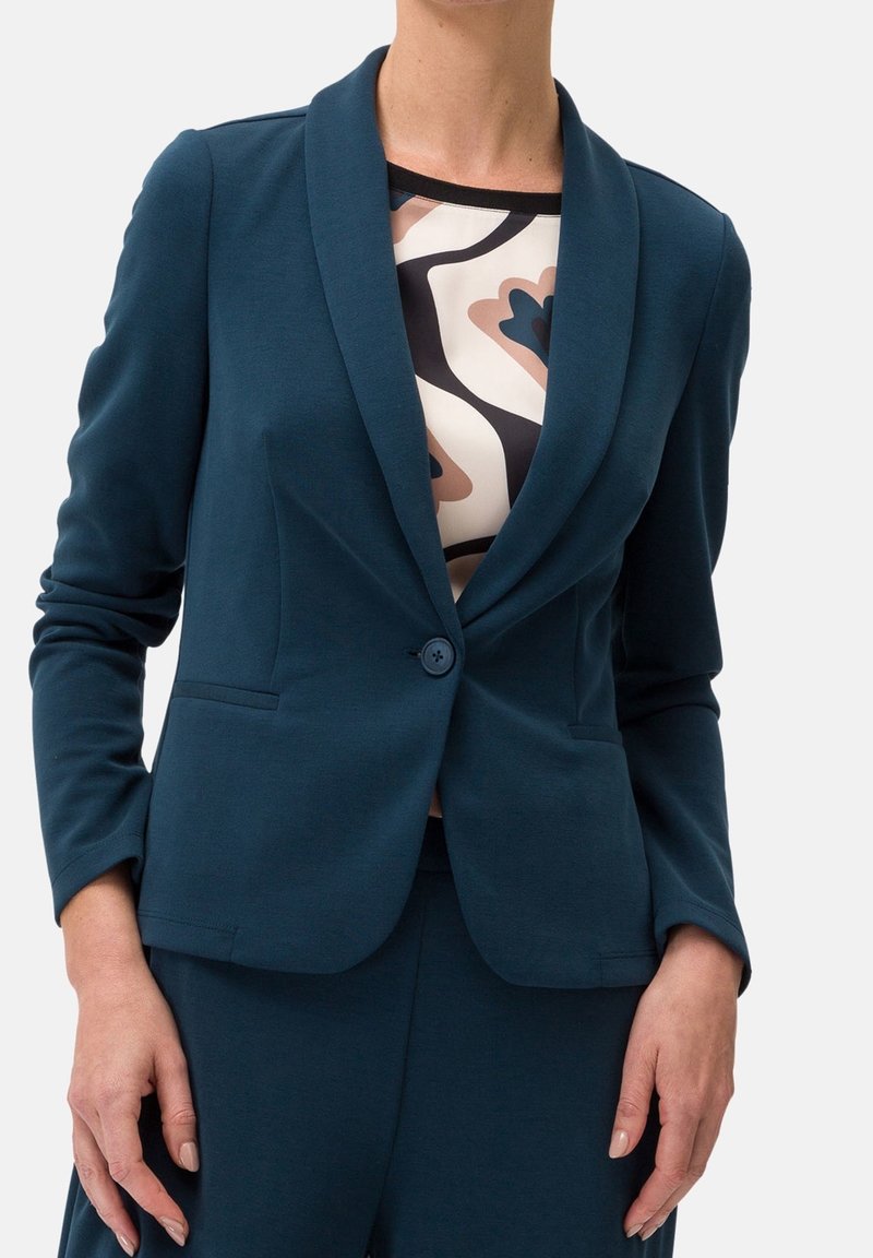 Blazer teal avec un col châle, fermeture à un bouton et deux poches avant. Porté par-dessus un haut à motifs aux couleurs beige, noir et teal.