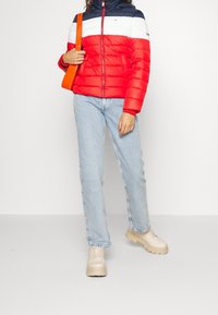 Kvinna iklädd en färgblockerad röd, vit och marinblå puffjacka, ljusblå jeans, beiga chunky stövlar och bär en ljusorange axelväska.