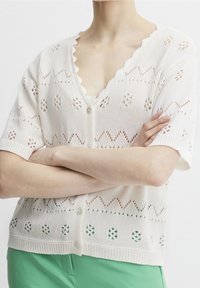 Person with arms crossed wearing a white short-sleeve knit cardigan with decorative holes and green pants, shown from shoulders to waist.