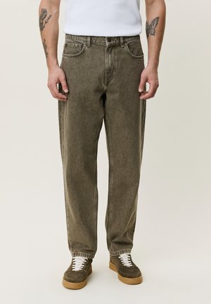 Mann trägt ausgewaschene olivgrüne Jeans, weiße Sneakers mit Gummisohlen und ein weißes Hemd, steht mit entspannt an den Seiten hängenden Armen.