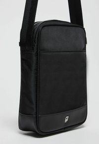 Borsa a spalla nera realizzata in tessuto resistente, con un design rettangolare elegante, una texture liscia e un piccolo logo argentato sulla parte anteriore.