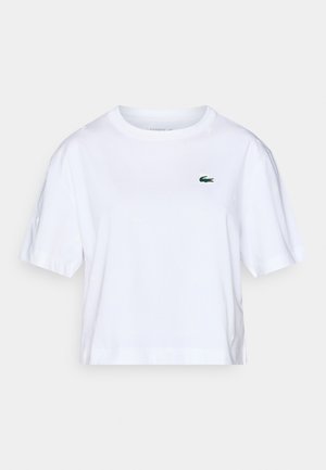 T-shirt court en coton blanc à manches courtes, avec un petit logo Lacoste vert sur la poitrine et un motif discret sur l'ensemble.