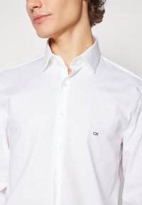 Ung person som bär en vit skjorta med knappar och en liten svart "ck"-logotyp på bröstet, med öppen krage och kort lockigt hår synligt.