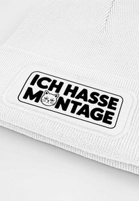 White knitted beanie featuring a rectangular patch with black text "ICH HASSE MONTAGE" and a cat illustration. Texture is ribbed.