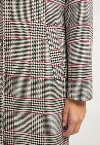Cappotto di lana con un motivo a quadri nero, bianco e rosso, tessuto testurizzato, una tasca e un dettaglio con bottone vicino al polsino.