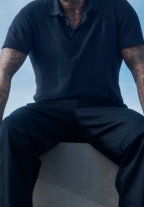 Homme aux bras tatoués assis portant un polo foncé et un pantalon devant un ciel bleu clair.