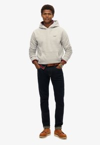 Sudadera gris con capucha y bolsillo frontal sobre una camisa de cuadros roja y azul, jeans oscuros y zapatos marrones con suelas claras. Conjunto casual.