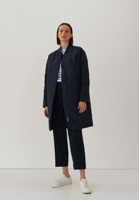 Marineblauwe geborduurde lange jas met een voorrits, enkele borstzak en geribde kraag, gecombineerd met een gestreept shirt en marineblauwe broek.