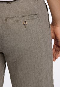 Pantalones en espiga marrones con un pequeño detalle de botón en el bolsillo trasero y una textura suave, que destacan un corte entallado y un patrón sutil.