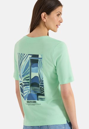 Femme portant un t-shirt vert menthe avec un motif abstrait de feuille bleue et fenêtre et le texte « SELFCARE » dans le dos, regardant par-dessus son épaule.