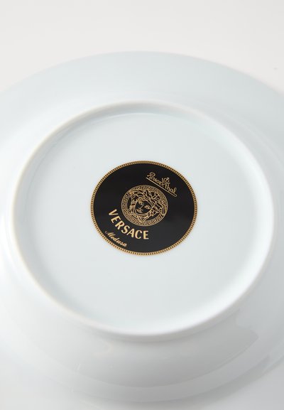 Prato de porcelana branca com um logotipo circular preto e dourado, apresentando o design Medusa da Versace no centro. Acabamento liso e brilhante.