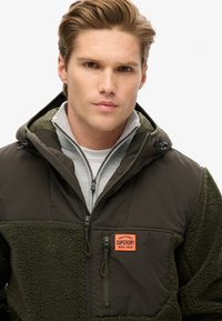 Giacca verde oliva con una parte inferiore in pile testurizzato, tasche con zip e una patch arancione. Caratterizzata da un cappuccio e un colletto grigio a strati.