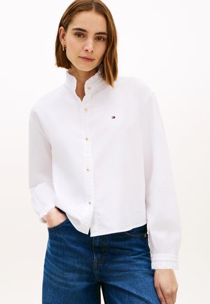 Kobieta o krótkich brązowych włosach, ubrana w białą koszulę na guziki ze złotymi guzikami i niebieskie dżinsy, stojąca z jedną ręką w kieszeni.