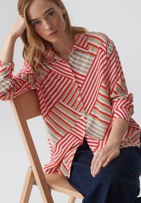 Camicia a righe rossa e beige con larghezze variabili, dotata di colletto e maniche arrotolate, indossata con pantaloni scuri.