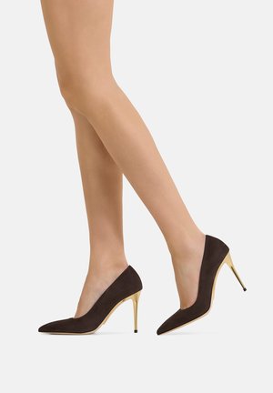 Gambe con décolleté nere a punta con tacco stiletto dorato, mostrati su uno sfondo grigio chiaro.