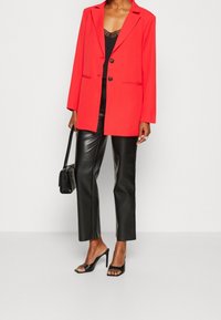Femme portant un blazer rouge vif sur un débardeur en dentelle noire, un pantalon en cuir noir, des sandales à talons noires, et tenant un petit sac à main noir.