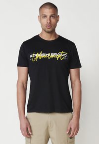 Camiseta de algodón negra con texto gráfico en blanco y amarillo, mangas cortas y escote redondo. Combinada con pantalones cortos de cargador beige.