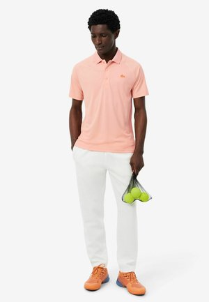 Uomo che indossa una polo rosa chiaro, pantaloni bianchi e scarpe da ginnastica arancioni che tiene in mano una borsa nera a rete con tre palline da tennis verdi.