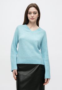 Mlada ženska v svetlomodrem puloverju z V-izrezom in črni usnjeni krilu, stoji pred preprostim belim ozadjem.
