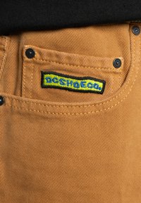Hnedé nohavice s textúrovaným povrchom, so sychrávkovým gombíkom na vrecku a vyšívaným logom "DC SHOE CO." v čiernej, modrej a žltej farbe.