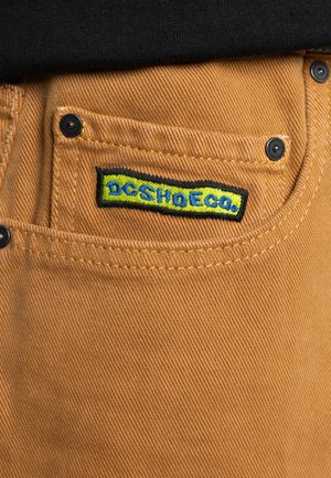 Helle Hosen mit strukturierter Oberfläche, ausgestattet mit einem dunklen Taschenschalter und einem schwarz-blau-gelben, gestickten "DC SHOE CO."-Logo.