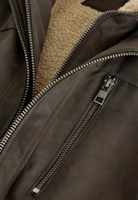 Braune Lederjacke mit einem Reißverschluss. Innen mit beigem, künstlichem Schaffell gefüttert. Sichtbare metallische Reißverschlussdetails auf der Vorderseite.