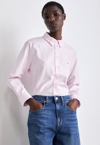 Camicia a righe rosa chiaro con colletto a punta; indossata con jeans blu. La camicia ha maniche lunghe e un piccolo logo sul petto.