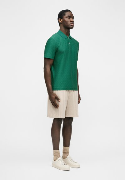 Άνδρας όρθιος, φοράει πράσινο πικέ πόλο πουκάμισο, μπεζ σορτς μέχρι το γόνατο, μπεζ κάλτσες και λευκά αθλητικά παπούτσια σε λευκό φόντο.