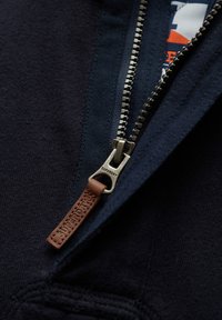 Donkerblauwe zip-up hoodie met een metalen rits en een leren trekband in bruin. Zachte stoftextuur met een casual ontwerp en gestikte details.