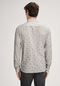 Man die wegkijkt en een lichtgrijze shirt met lange mouwen en een geometrisch patroon draagt, samen met donkere jeans tegen een effen achtergrond.