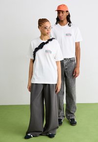 Two individuals wear casual attire: a white T-shirt with colorful text, gray wide-leg pants, and light gray denim jeans. Green flooring.