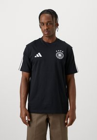 Νεαρός άνδρας με πλεξίδες, φορώντας μαύρο μπλουζάκι Adidas με το λογότυπο της Γερμανικής Ομοσπονδίας Ποδοσφαίρου και τέσσερις αστέρες, που στέκεται μπροστά σε λευκό φόντο.