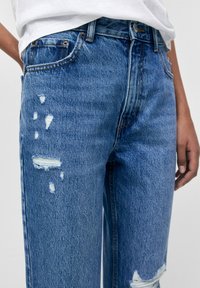 Vaqueros de mezclilla azul desgastados con un corte relajado, que presentan deshilachados ligeros y varios parches rasgados en la parte delantera. Diseño clásico de cinco bolsillos.