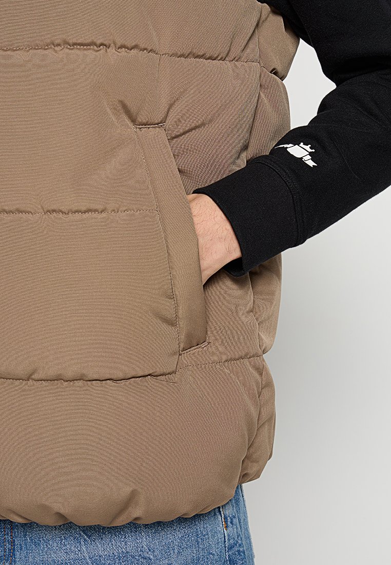 Weste aus leichtem braunem Material, aus gestepptem Stoff. Verfügt über Reißverschlusstaschen an den Seiten und ein schwarzes Langarmshirt darunter mit Logodetail.