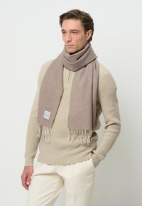 Sciarpa di lana beige con estremità frangiate, appoggiata attorno al collo, con un piccolo etichetta sul lato. Il modello indossa un maglione crema e pantaloni chiari.