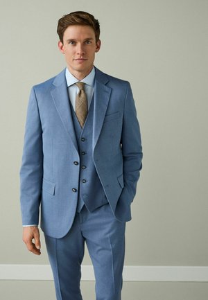 Mand iført lyseblå tredelt jakkesæt med lyseblå skjorte og mønstret brun slips, stående foran en ensfarvet grå væg.