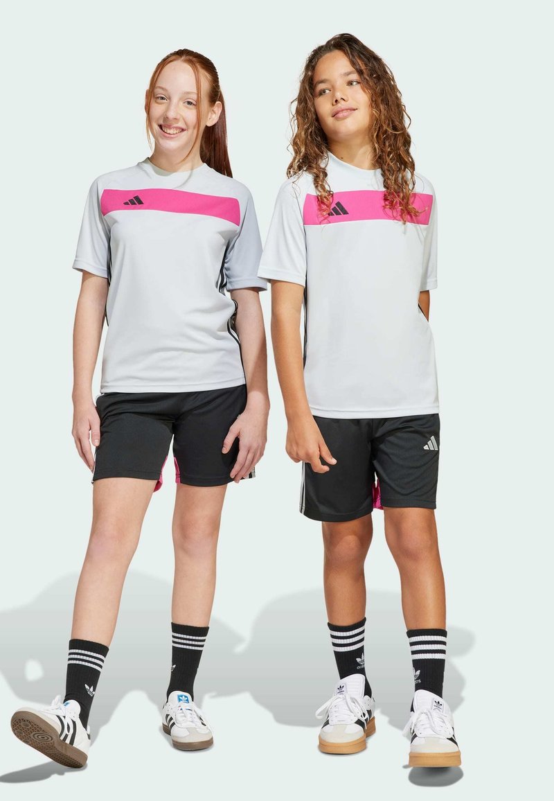 Dos niños con camisetas deportivas grises con rayas rosas, pantalones cortos negros y calcetas negras con rayas blancas, usando calzado Adidas.