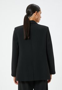 Blazer nero su misura con maniche lunghe, apertura sul retro e design delle spalle strutturato. Tessuto liscio e opaco con dettagli minimi.
