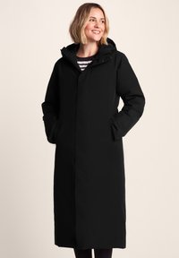 Woman smiling, wearing a long black hooded winter coat with hands in pockets, standing against a plain light background.